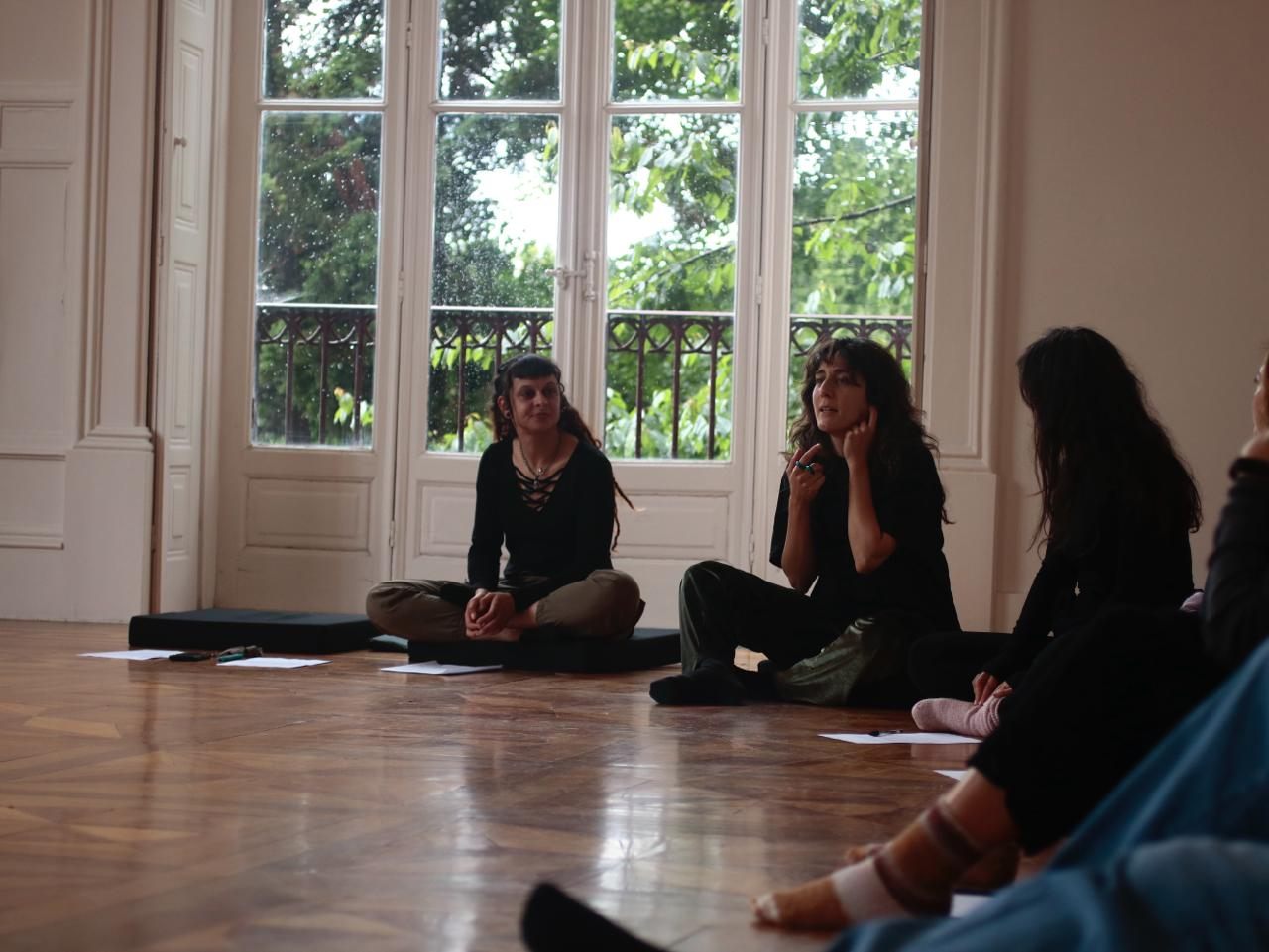Dila in conversation with participants, seated on the floor before the windows