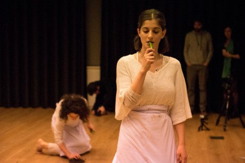 A performer in white standing with a hand at her mouth, another kneeling behind her