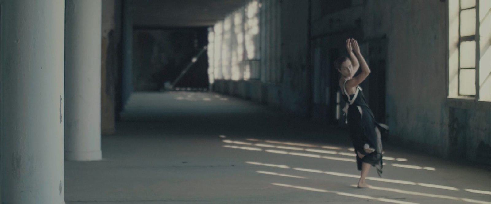 A dancer in black in a derelict hall lit by tall industrial windows
